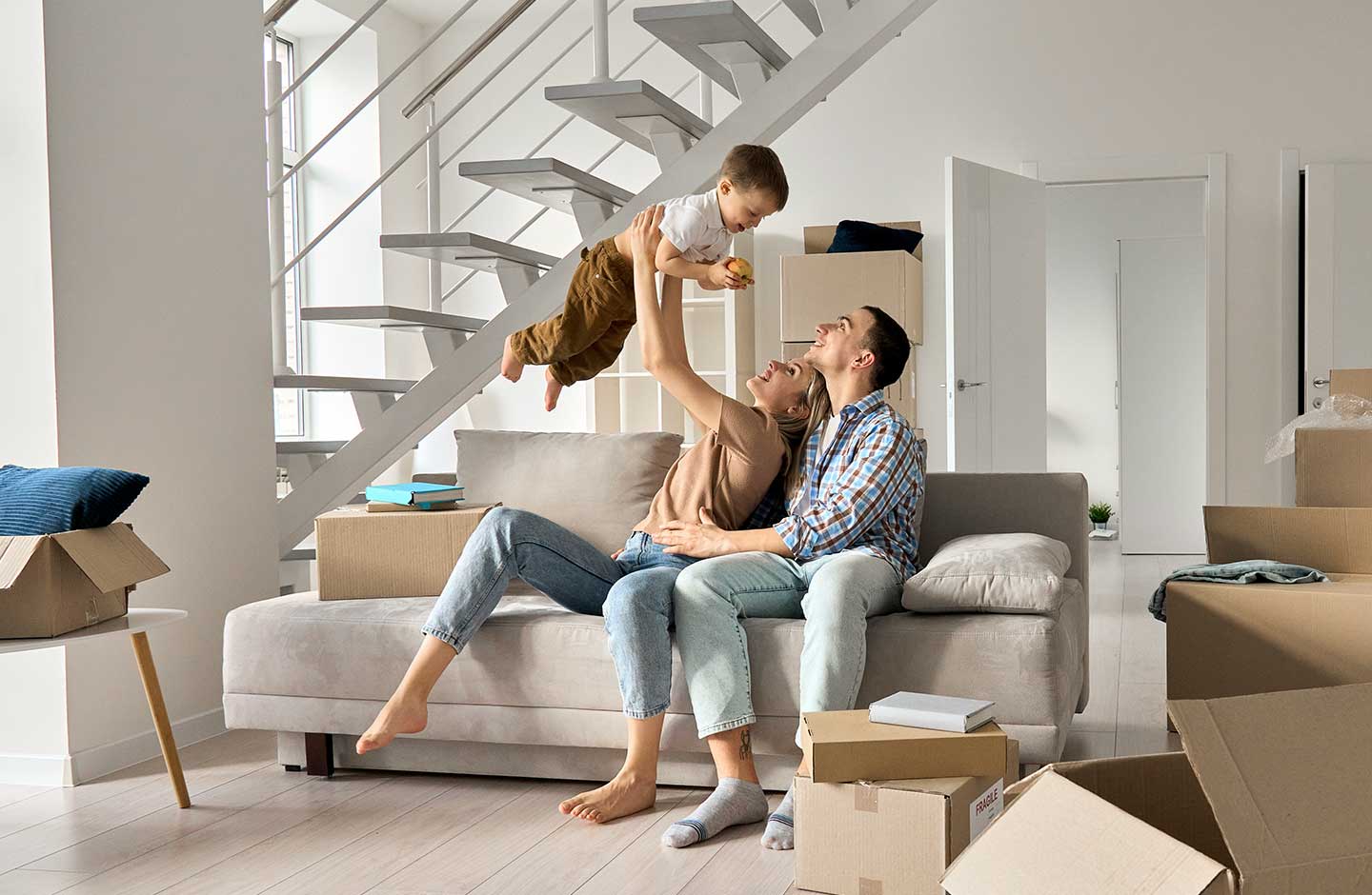 Mutter hebt Kind in die Luft, Familie sitzt auf dem Sofa in ihrer neuen Wohnung