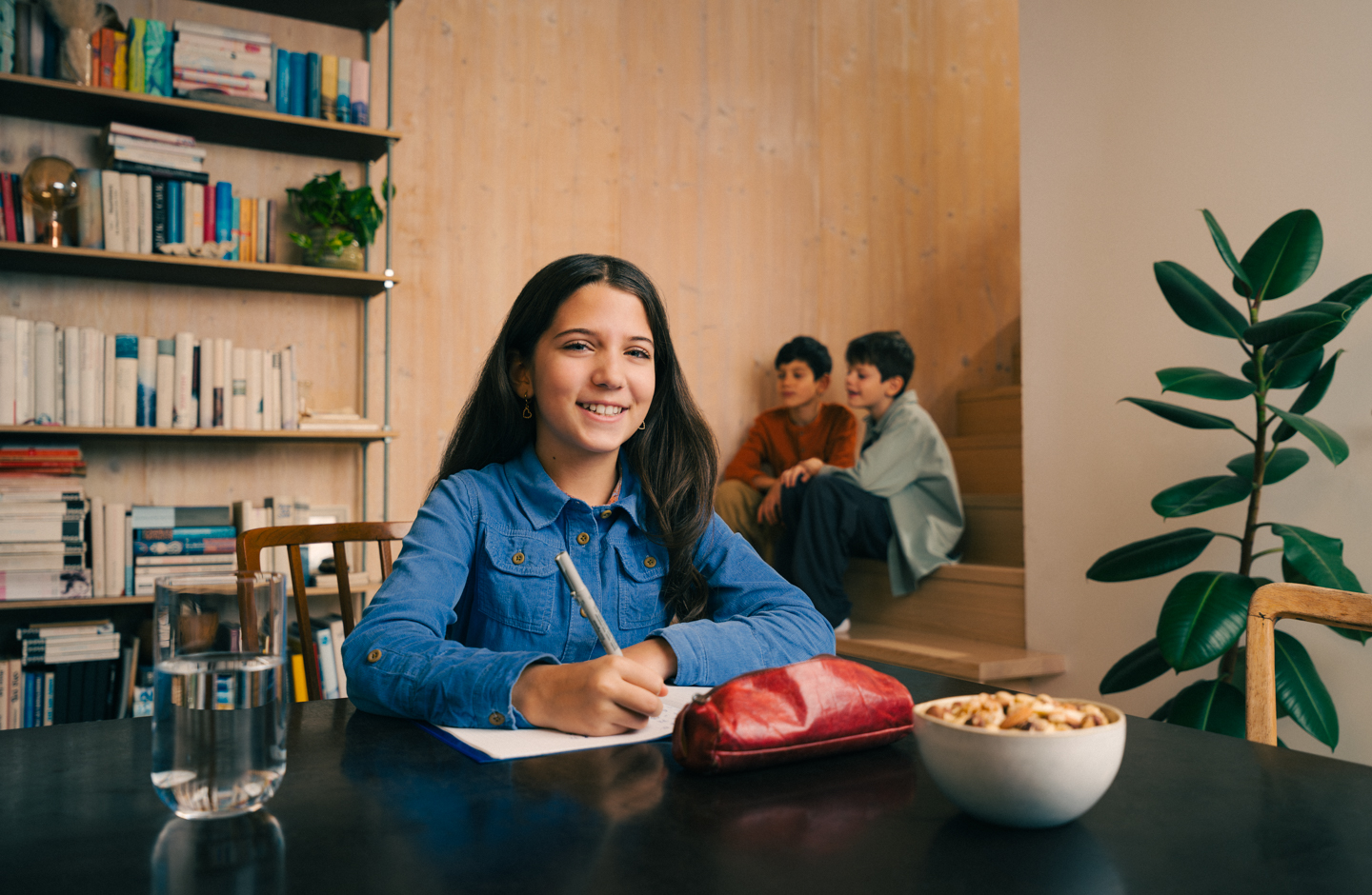 Ein Mädchen sitzt an einem Tisch bei den Hausaufgaben. Sie hält einen Stift in der Hand und lächelt in die Kamera.