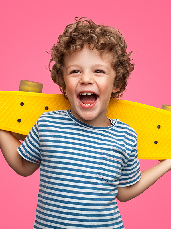 Kleiner Junge lacht mit gelben Skateborad auf dem Ruecken und vor einem rosa Hintergrund