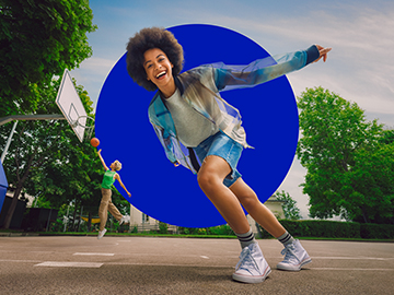 Zwei junge Frauen spielen Basketball. Eine der Frauen wirft im Springen einen Korb, die andere läuft an der Kamera vorbei und lacht.