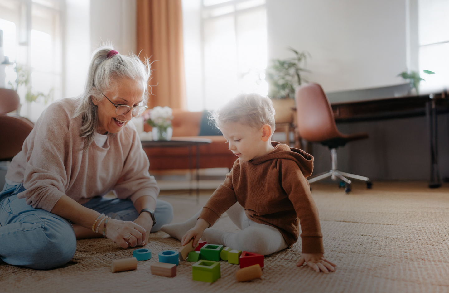 Eine ältere Frau sitzt mit einem Kind auf dem Fußboden. Sie spielen mit Holzspielzeug.