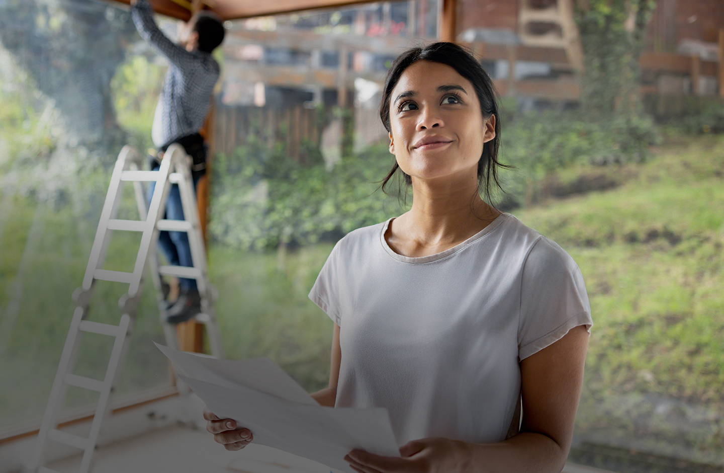 Eine junge Frau steht in ihrem neuen Haus, das gerade gebaut wird. Sie hat einen Bauplan in der Hand.